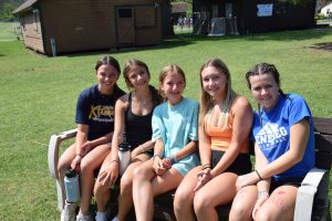 5 female campers sitting on a bench in the grass smiling at the camera.