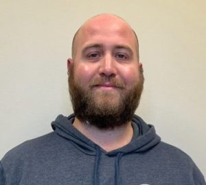 Bald man with a beard and mustache posing for the camera with a gray hooded sweatshirt on.